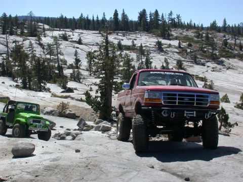 1996 Ford Bronco - Two off-road vehicles navigate a rocky, wooded landscape under a clear blue sky.