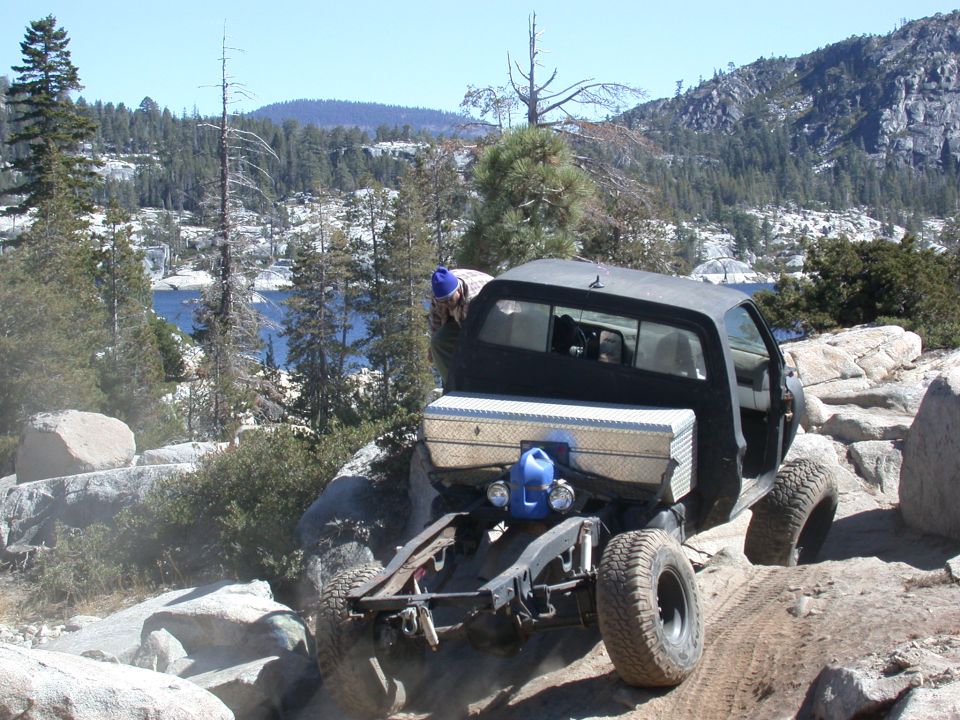 Pickup truck with a toolbox and gas can on the back, driving over rocks near a lake