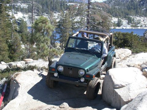 1996 Ford Bronco - A green Jeep Wrangler navigates a rocky trail with a lake in the background.