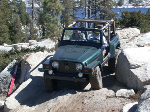 1996 Ford Bronco - A green Jeep Wrangler tackles a rocky trail with a lake and mountains in the background.