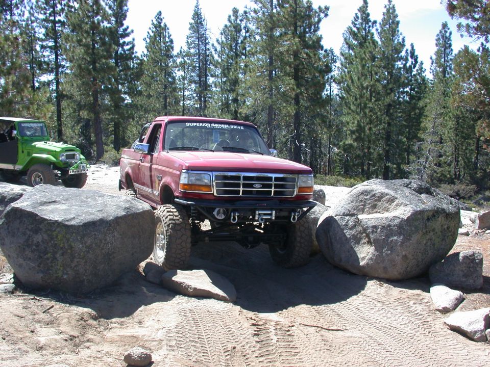 Red Ford F-Series truck with large tires navigating rocks, green Jeep in background