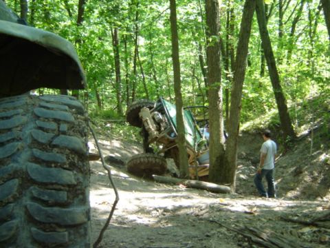 1992 Ford Bronco - An off-road vehicle is tipped on its side in a forest, with a person standing nearby and another vehicle's tire visible.