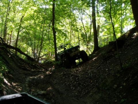 1992 Ford Bronco - An off-road vehicle navigates a steep, muddy trail surrounded by dense trees.