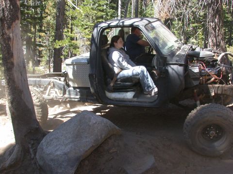 1996 Ford Bronco - Two people ride in a modified off-road truck with its engine exposed, navigating a wooded trail.