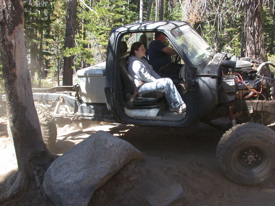 Off-road truck with two people in the cab, engine exposed, driving on dirt trail