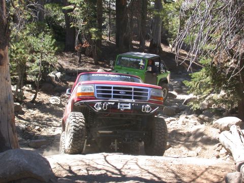 1996 Ford Bronco - Two off-road vehicles navigate a challenging, rocky path through a wooded area.
