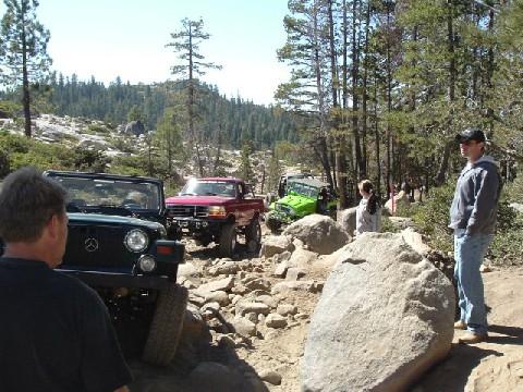 1996 Ford Bronco - Several off-road vehicles navigate a challenging rocky trail through a wooded area.