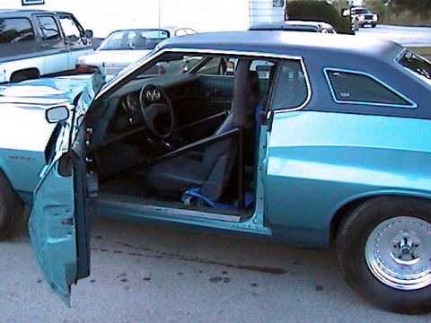 1977 Ford F-150 - The interior of a two-tone blue Ford Gran Torino Sport shows a roll cage and racing seat, with other vehicles in the background.