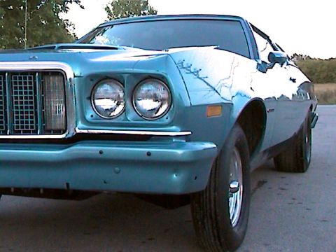 1977 Ford F-150 - The front end of a blue 1977 Ford Gran Torino is visible from a low angle, highlighting its distinctive grille and headlights.