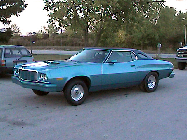 Blue 1977 Ford Gran Torino coupe with black vinyl top parked outside, another vehicle to the left and a truck to the right