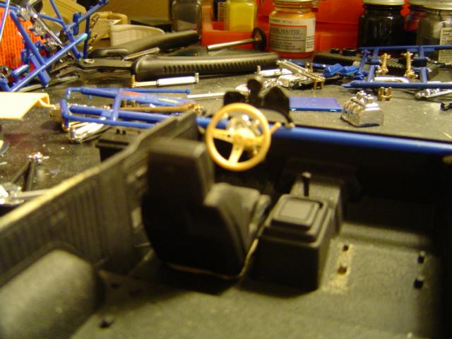Model car interior with black seats, yellow steering wheel, and a gear shift. Parts and tools are on the workbench