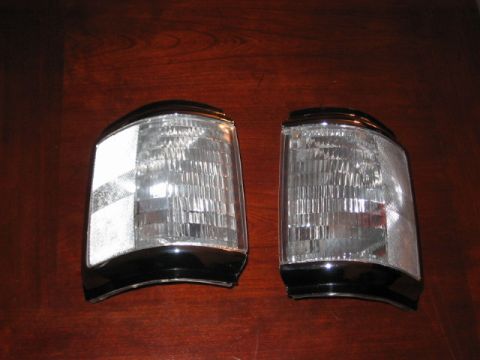 1991 Ford Bronco - A pair of vehicle headlights with clear lenses and black trim are displayed on a wooden surface.