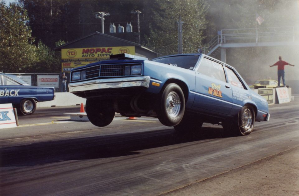 Blue drag car doing a wheelie on a track, with another car visible to the left