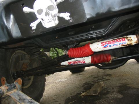 1985 Ford Bronco - Undercarriage view shows Skyjacker Hydro 7000 shocks and red boots, with a skull and crossbones sticker mounted above.