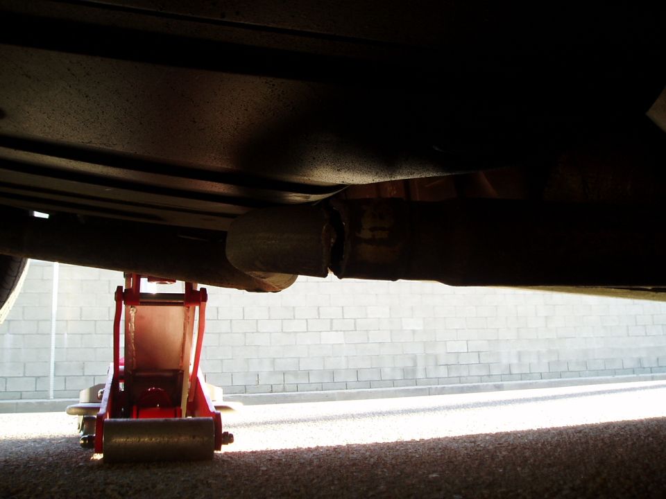 Underside of a vehicle with a red jack supporting the frame near a broken exhaust pipe