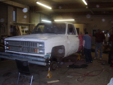 1981 Chevrolet C/K Pickup - A white pickup truck with a red bed is undergoing repairs in a garage, supported by jack stands.