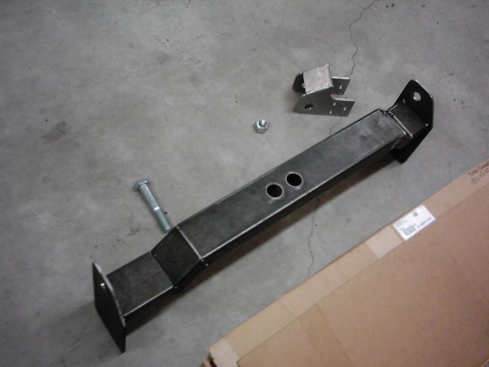 Metal frame part with two circular holes, a bolt, and a nut on a concrete floor next to a cardboard box