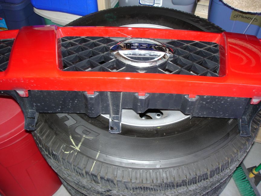 Red Nissan grille with chrome emblem and bottom mounting clips, resting on a tire.