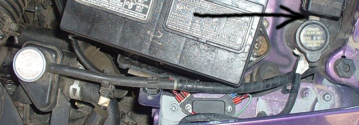 Engine bay close-up with arrow pointing to coolant overflow bottle cap beside the battery