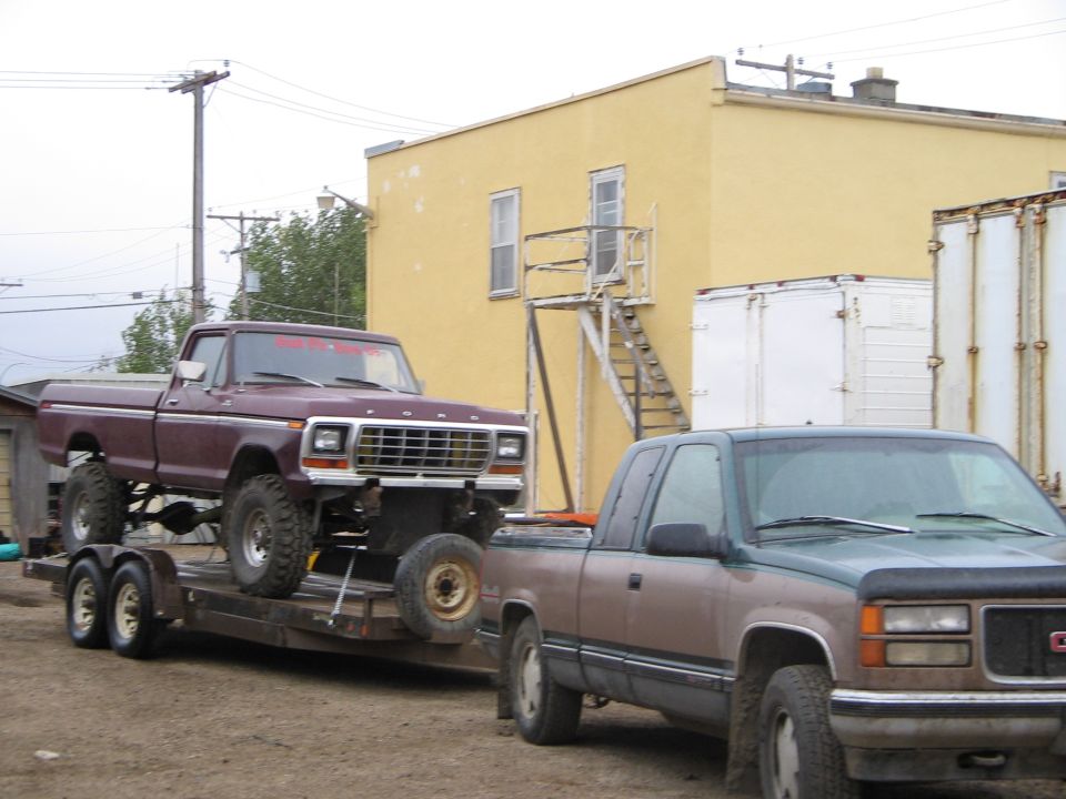 Maroon Ford F-Series pickup on trailer, towed by GMC pickup. Yellow building in background