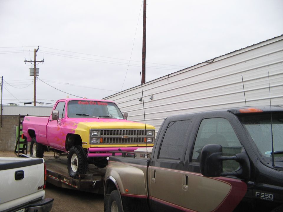 Pink and yellow pickup truck on a trailer, being towed by another truck. Text on windshield says 'Push It To The Limit'
