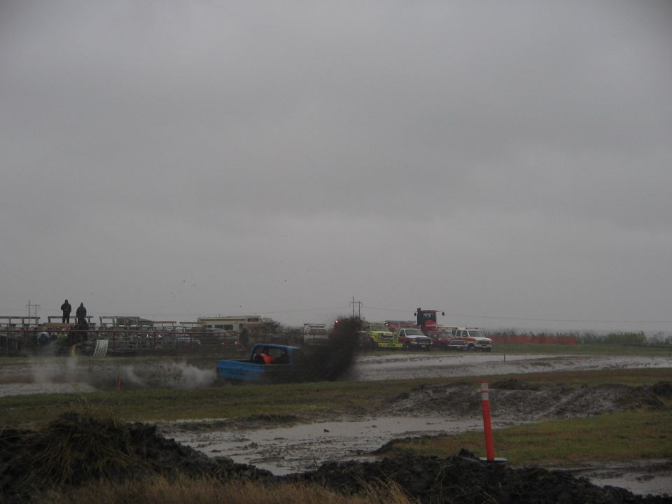 Blue pickup truck kicking up mud during a race, with other vehicles and spectators in the background under a cloudy sky