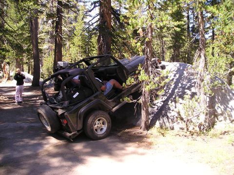 1989 Ford Bronco - An off-road vehicle is stuck on a large rock with trees on either side, while a person stands nearby.