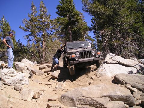 1989 Ford Bronco - A person pushes a Jeep Wrangler through a challenging rocky trail on a sunny day.