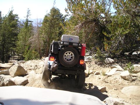 1989 Ford Bronco - A Jeep navigates a rocky off-road trail with trees and boulders surrounding it.