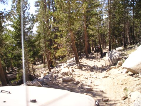 1989 Ford Bronco - A trail winds through a rocky, wooded area, with two vehicles navigating the terrain in the distance.