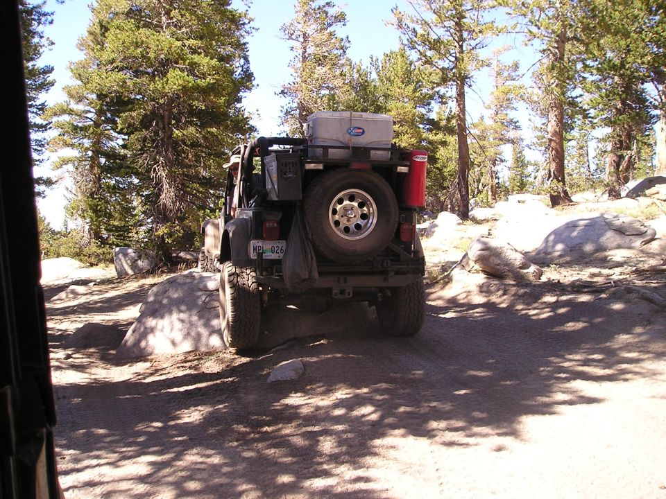 Rear view of a Jeep Wrangler with a spare tire, cooler, and red canister on the back, driving on a dirt trail