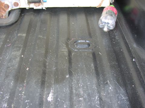 1979 Ford Bronco - The interior of a truck bed shows a ribbed floor with a tie-down anchor and a discarded plastic bottle.