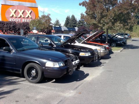 1999 Ford Crown Victoria - Several Ford Crown Victorias are parked in a lot with their hoods up, near a XXX Root Beer sign.