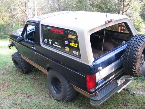 1995 Ford Bronco - This lifted Ford Bronco sits parked on grassy terrain, showing off its off-road modifications and various stickers on the rear window.