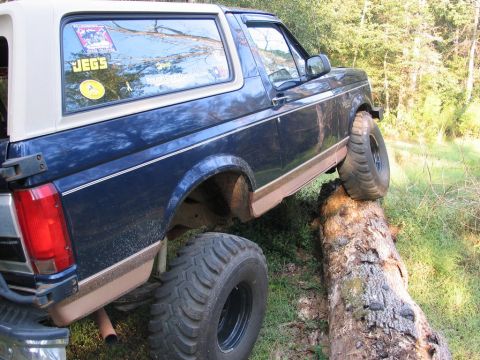 1995 Ford Bronco - A blue Ford Bronco navigates an obstacle course, driving over a fallen tree in a wooded area.