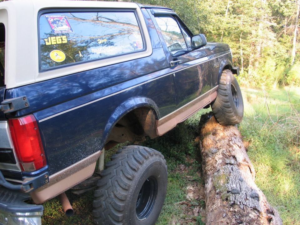 Blue Ford Bronco with a white top, off-roading over a large log. Mud splatters on the lower body