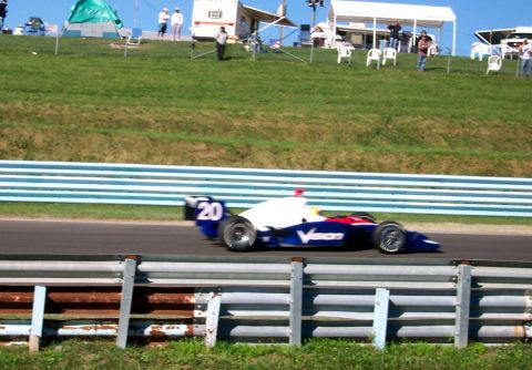 1979 Ford F-150 - A race car speeds around a track with spectators and tents visible on the grassy hillside in the background.