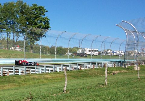 1979 Ford F-150 - An open-wheel race car speeds along a track, with RVs visible in the spectator area beyond the fence.