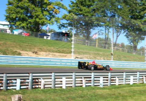 1979 Ford F-150 - A race car speeds around a track on a sunny day, with trees and tents in the background.