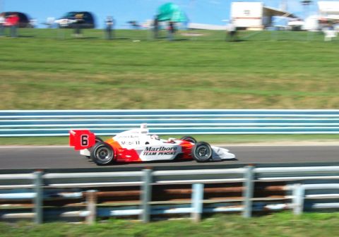 1979 Ford F-150 - A race car speeds along the track, its red and white livery clearly visible against the green hillside.