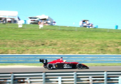 1979 Ford F-150 - A red and black open-wheel race car speeds around a track on a sunny day.