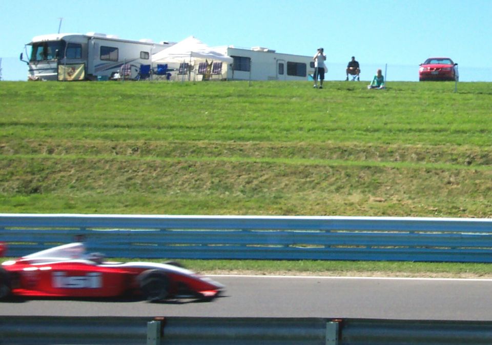 Red open-wheel race car on track, RVs and people on grassy hill in background