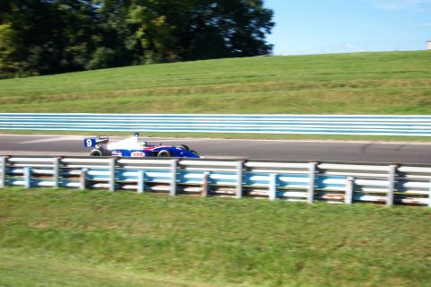 1979 Ford F-150 - A blue and white open-wheel race car speeds along a track, with grassy hills and blue and white barriers in the background.