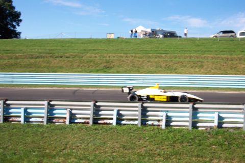 1979 Ford F-150 - A race car drives on a track with a grassy hill and blue and white barriers in the foreground.