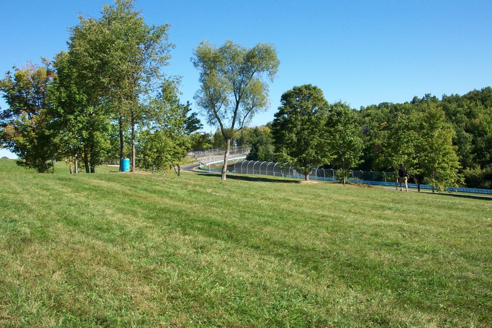 Grassy hill with trees and a blue barrel under a clear blue sky. A fence and road are visible in the background