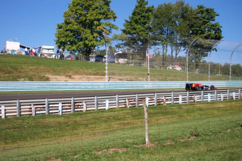 1979 Ford F-150 - A single-seater race car speeds along the track with spectators and trees in the background.
