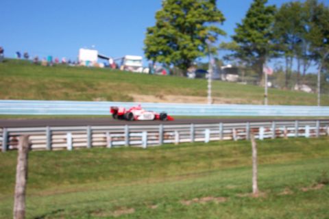 1979 Ford F-150 - Race car speeds around a track on a clear day with trees and spectators visible.