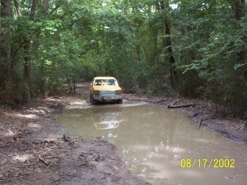 1991 Ford Bronco - Yellow SUV navigates a muddy trail and puddle surrounded by dense trees. The image is dated August 17, 2002.