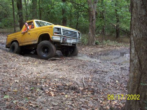 1991 Ford Bronco - A yellow Ford F-Series truck navigates a muddy trail in a wooded area.