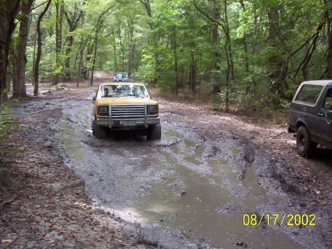 1991 Ford Bronco - A yellow SUV tackles a challenging muddy path in a wooded area, with another vehicle visible in the distance and a dark SUV parked on the s…
